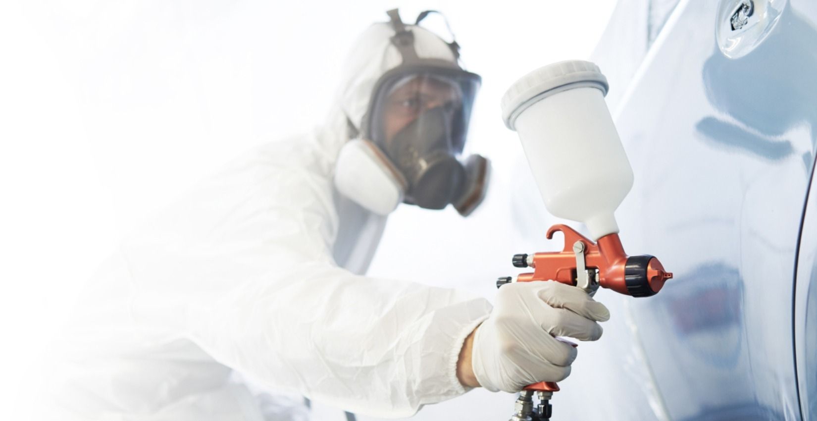 Person in Schutzanzug und Atemmaske hält eine Spritzpistole und lackiert eine Oberfläche
