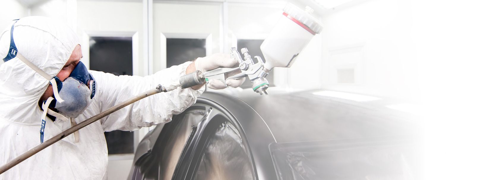 Person in Schutzanzug und Atemmaske sprüht Lack auf eine Fahrzeugkarosserie
